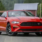 Red Ford Mustang convertible seen from the front-left, parked on a rural road with trees and a white fence in the background; headlights on.