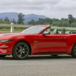 Red Ford Mustang convertible with top down, parked on a rural road beside a white fence and hills in the distance.