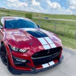 Red Ford Mustang with white racing stripes parked on a paved road beside a chain-link fence and grassy field under a blue, partly cloudy sky.