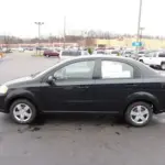 Black four-door sedan parked in a car dealership lot with a price sheet visible on the window.