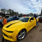 Yellow sports car with a black racing stripe at an outdoor car show, flag in the window and people in the background.