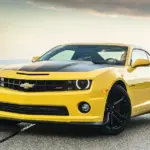 Front three-quarter view of a bright yellow Chevrolet Camaro with a black hood, parked on a paved road at sunset, wheels turned slightly to the right.