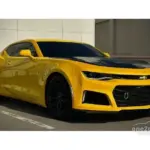 Yellow Chevrolet Camaro with a black hood and black wheels parked in an outdoor lot.