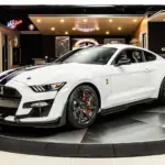 White Ford Mustang Shelby GT500 on a circular showroom turntable with glossy black-and-white floor and neon signs in the background, angled to show the car's front grille and logo.