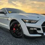 White Ford Mustang Shelby GT500 in a front three-quarter view, parked on a rural road at sunset; black wheels with red brake calipers.