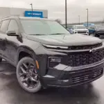 Black Chevrolet SUV parked on a wet dealership lot, front view showing the distinctive grille and LED headlights, with other cars and a Chevrolet sign in the background.