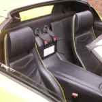 Open-top sports car cockpit with black leather seats, yellow piping, and racing harnesses
