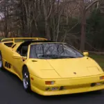 Yellow sports car with the roof down parked on a paved road beside a wooded area behind it.
