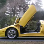Yellow sports car with doors raised upward (scissor/gull-wing style) parked on a driveway, stone wall and trees in the background