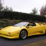 Bright yellow convertible sports car with doors open, parked on a driveway beside a stone wall and green hedges.