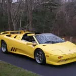 Yellow Lamborghini Diablo roadster parked on a paved road with leafless trees in the background, the top down and rear spoiler visible.
