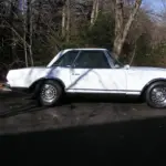 Classic white vintage car with chrome wheels parked on a paved lot, bare trees in the background.