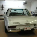 Rear of a white vintage Mercedes-Benz 280SL with automatic transmission parked in a cluttered garage; Massachusetts plate 26WF94 visible, fishing rods on wall left and ladder tools on the right.