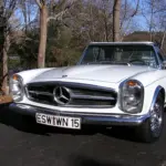 Front three-quarter view of a white classic Mercedes convertible parked on a paved surface with leafless trees in the background, winter scene.