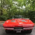 Front view of a bright red vintage convertible in a driveway, with the top down and green trees in the background.