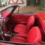 Red vintage convertible interior with red leather seats and open top, parked outdoors near a white barn door.